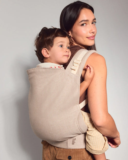A woman wearing a beige tula toddler carrier with a young child in a side-by-side baby carrier, shown against a plain light gray studio background.
