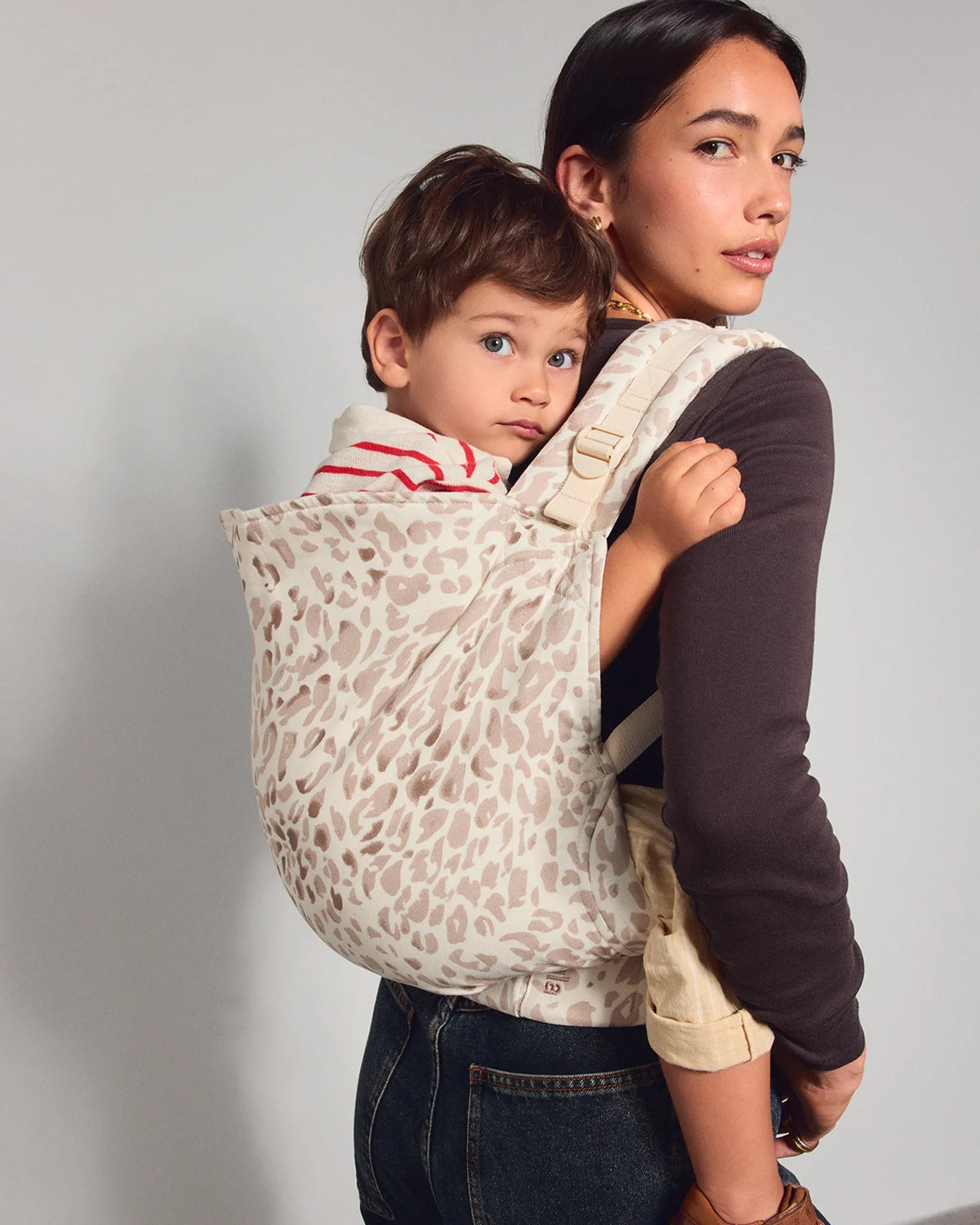 A woman wearing a leopard-print baby carrier with a young child inside, shown from the back against a plain light gray studio background.