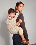 A woman wearing a leopard-print toddler carrier with a young child on her back, photographed against a plain light gray wall.