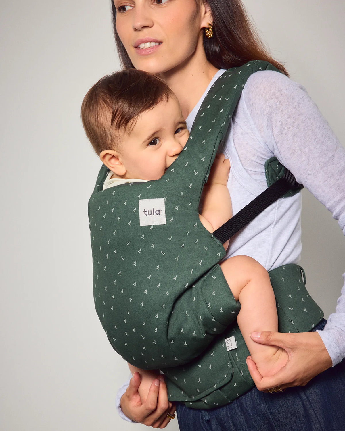 A woman wearing a Tula explore baby carrier with a small white pine tree pattern on a dark green background, holding a baby against her chest.
