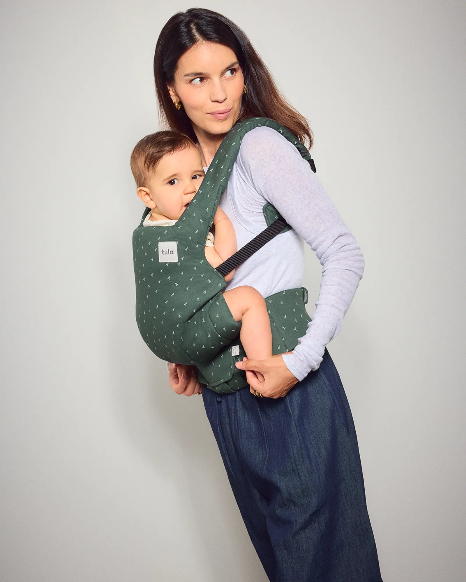 A woman wearing a dark green explore baby carrier with white polka dots carries a baby against her chest. The baby is wearing a white shirt and blue pants.