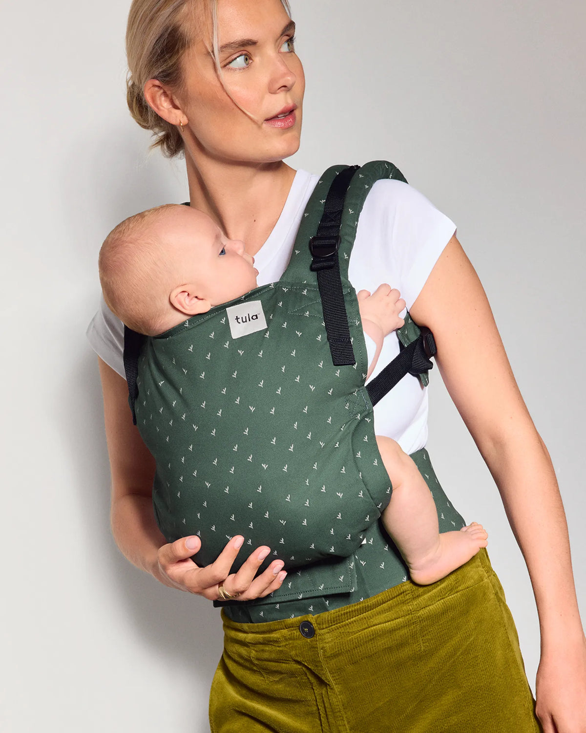A woman wearing a green original baby carrier with a small white leaf pattern, holding a baby against her chest. She is wearing a white t-shirt and olive green corduroy pants, photographed against a neutral light gray background.