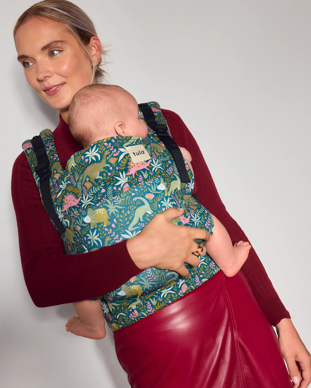A woman wearing a burgundy long-sleeve top and a red faux-leather skirt carries a baby in a green patterned tula original (formerly free-to-grow) baby carrier, standing in a minimalist white room corner.