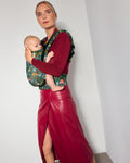 A woman wearing a burgundy long-sleeve top and red faux-leather skirt carries a baby in the tula original (formerly free-to-grow) baby carrier. The carrier has a colorful tropical print with green, pink, and yellow dinosaurs and plants on a teal background.