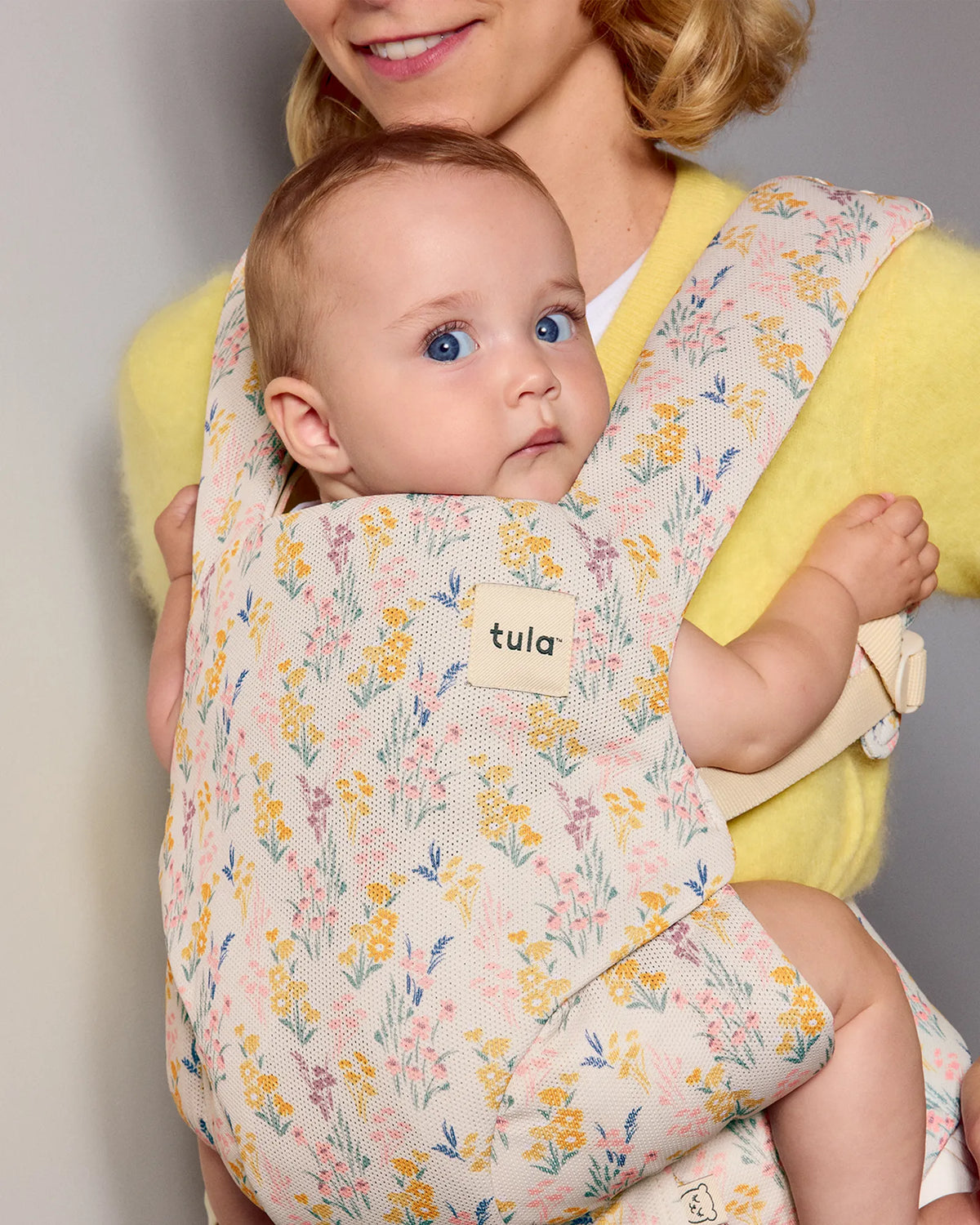 A tula explore baby carrier with a pastel floral print worn by a smiling woman holding a baby. The carrier has a cream base with yellow, pink, and blue flowers.