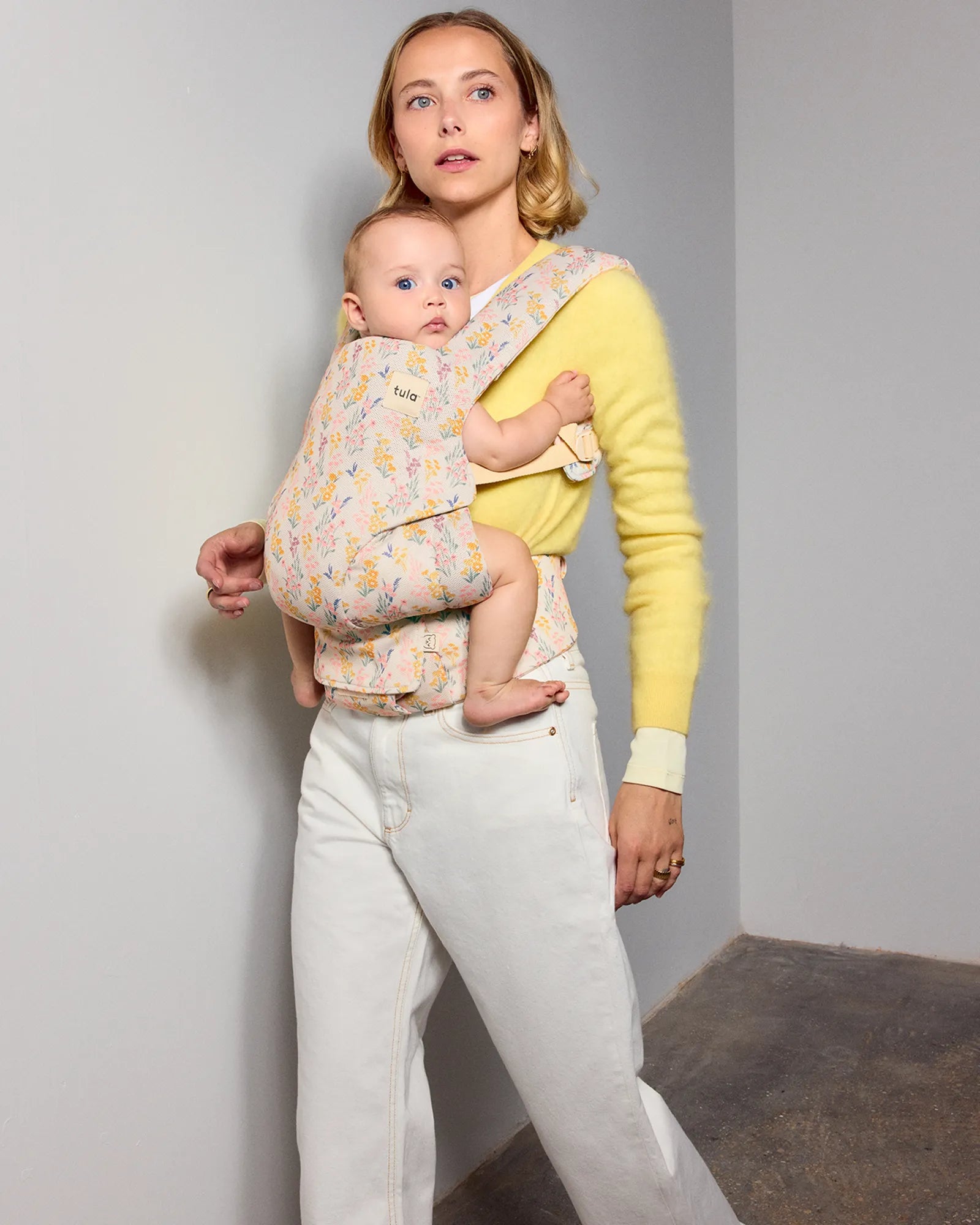 A woman wearing a yellow sweater and white pants carries her baby in a light pink floral tula explore baby carrier against a plain gray wall.
