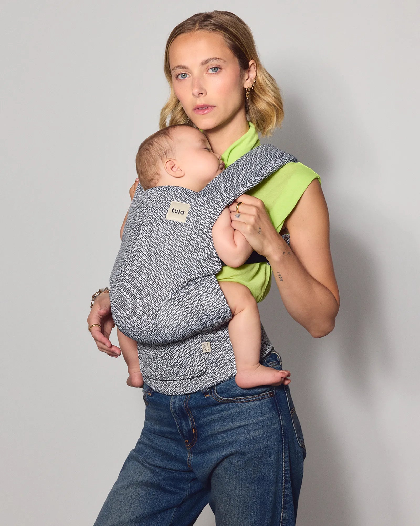 A woman wearing a gray patterned tula explore baby carrier with a baby inside, standing against a light gray studio background. 