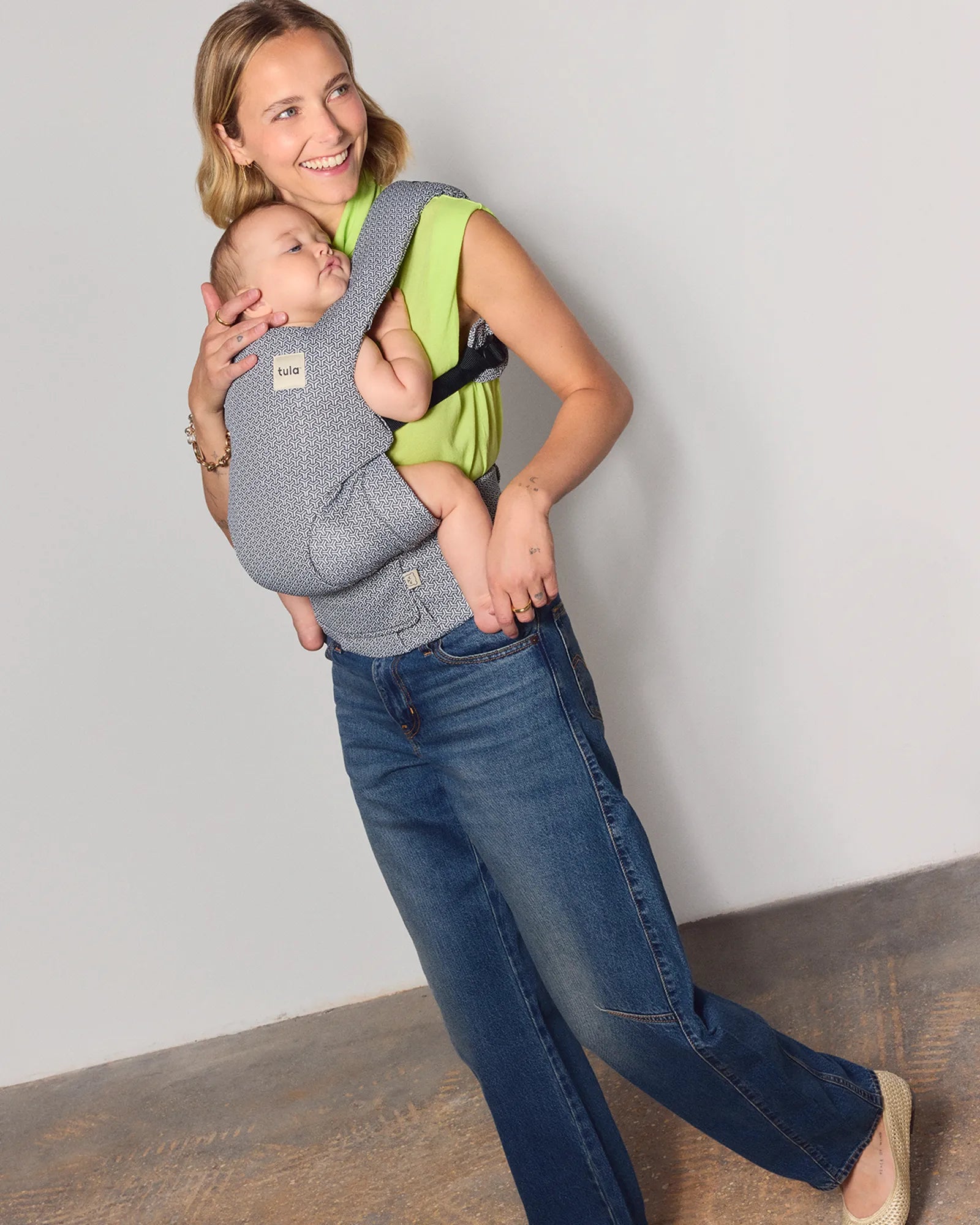 A woman wearing a bright lime green tank top and blue jeans carries a sleeping baby in a gray patterned tula explore baby carrier wrap. The woman is standing in a studio with a light gray wall and a textured brown floor.