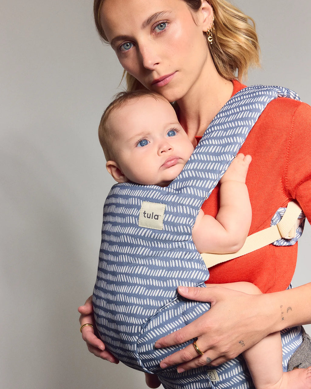 A woman wearing a Tula explore baby carrier with a blue and white wavy pattern and a bright orange-red inner wrap holding a baby, photographed against a neutral gray studio background.