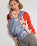 A woman wearing a red long-sleeve top and gray trousers carries a baby in a blue and white patterned Tula explore baby carrier against a plain light gray background.