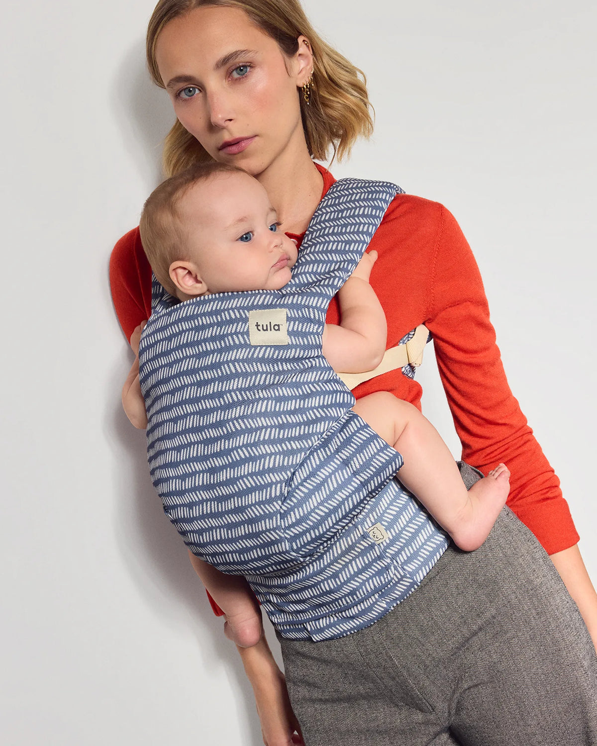 A woman wearing a red long-sleeve top and gray trousers carries a baby in a blue and white patterned Tula explore baby carrier against a plain light gray background.