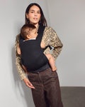 A woman wearing a leopard-print top and brown wide-leg trousers carries a baby in a black tula explore baby carrier. She is standing in a minimalist studio with a light gray wall and concrete floor.