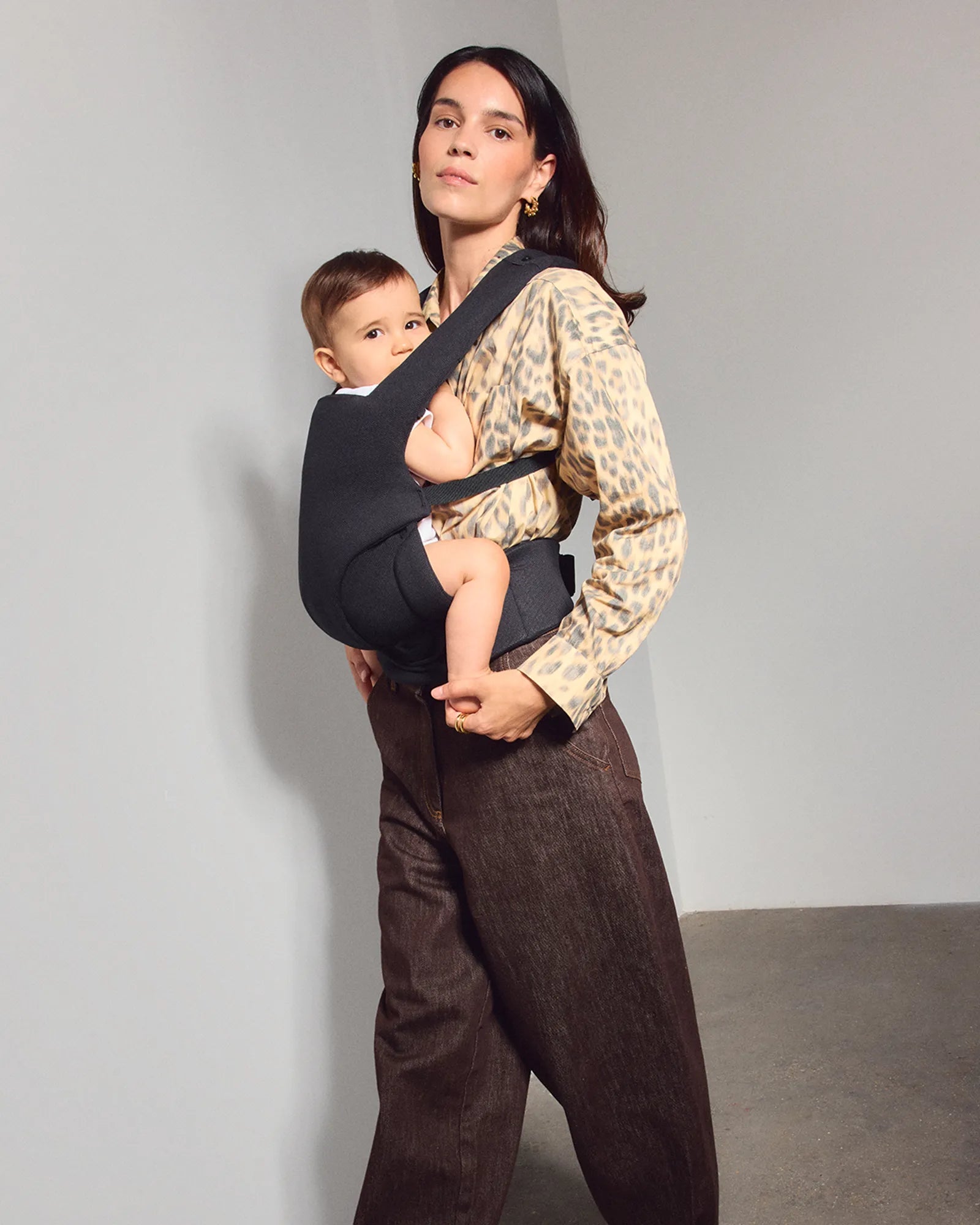 A woman wearing a leopard-print shirt and brown pants carries a baby in a black front tula explore baby carrier against her chest, standing against a plain light gray studio wall.