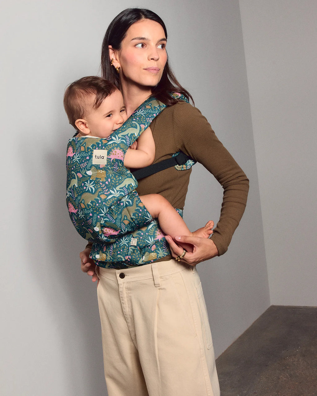 A woman wearing a brown long-sleeve top and beige pants carries a baby in a floral baby carrier wrap. The baby wears a matching patterned Tula Explore baby carrier with a blue, green, pink, and yellow tropical-style print.