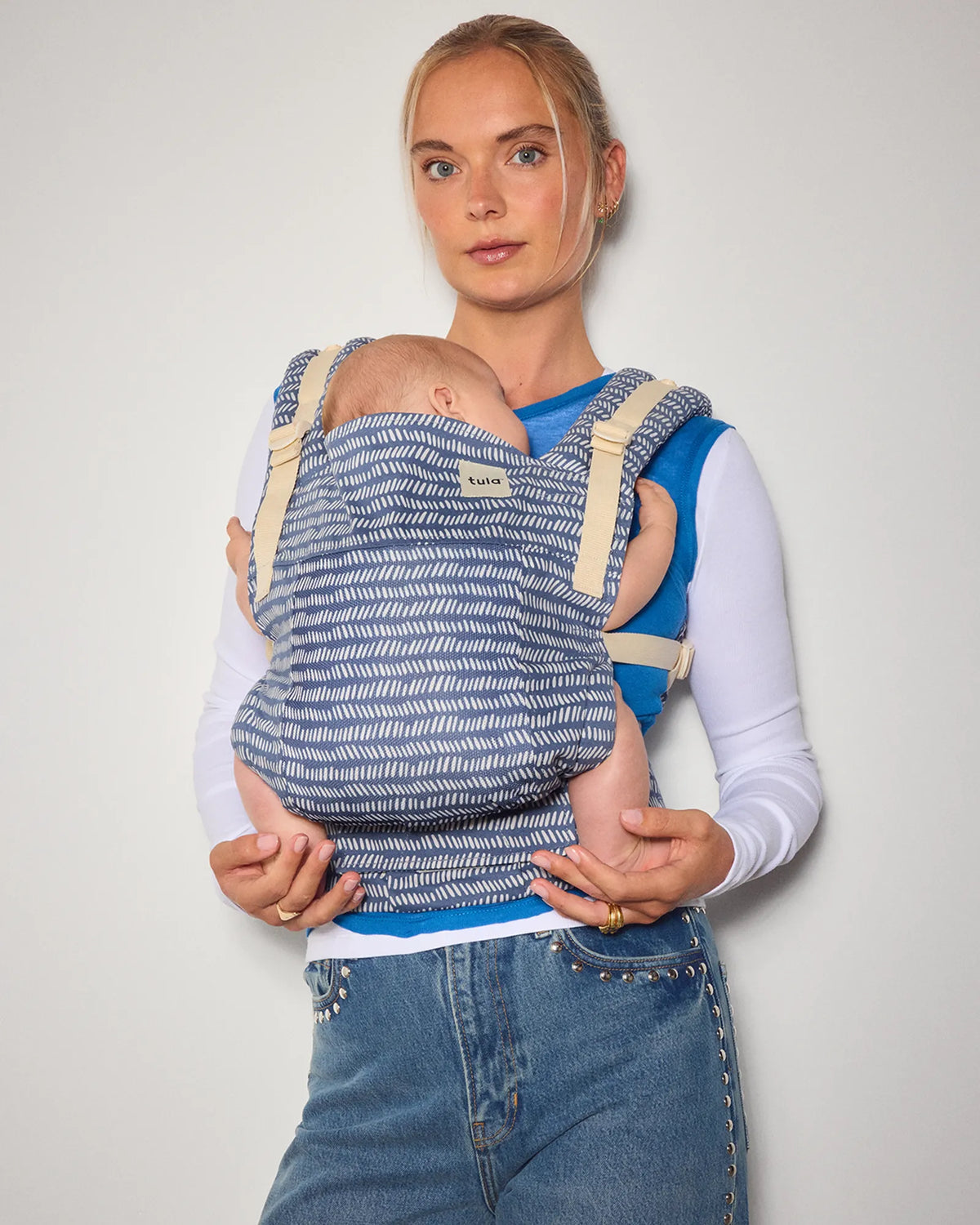 A woman wearing a blue and white striped original (formerly free-to-grow) baby carrier with a baby inside, standing against a plain light gray background.