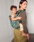 A woman wearing olive green pants and a brown ribbed long-sleeve top carries a young child on her back using a floral jungle-print toddler carrier. The carrier features a green background with pink, blue, and yellow tropical animal and plant illustrations.