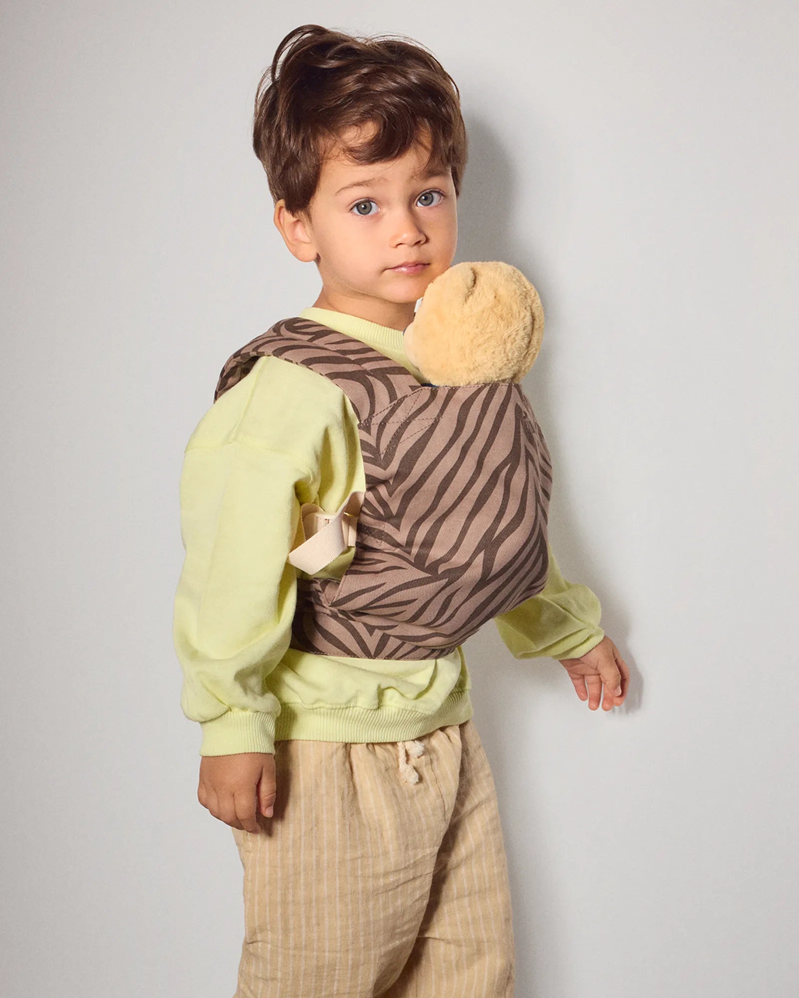 A toddler standing in a studio wearing a brown and beige zebra-print mini baby carrier wrap over a pale yellow-green shirt and beige pants, with a plush tan toy tucked into the carrier.