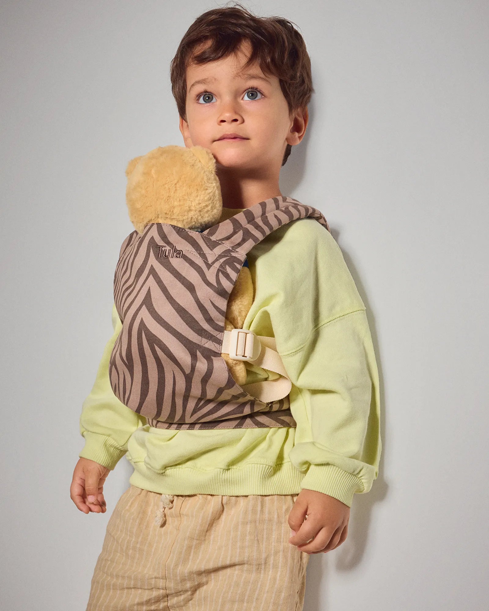 A young child wearing a light green sweatshirt and beige striped pants, shown from the waist up in a baby carrier with a brown and beige zebra-patterned fabric and a beige plush insert, against a plain light gray background.