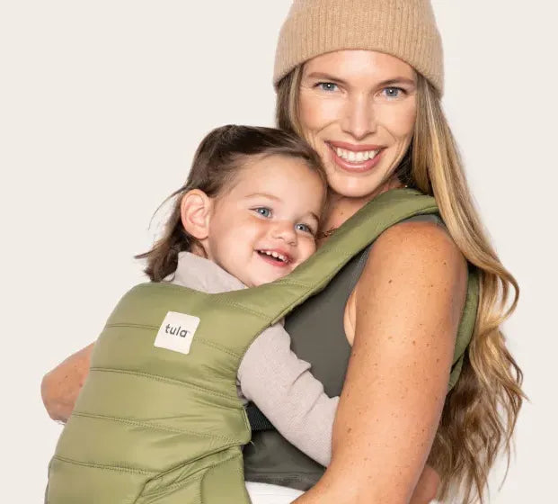 Woman carrying a child in a green Tula puffer baby carrier against a plain background