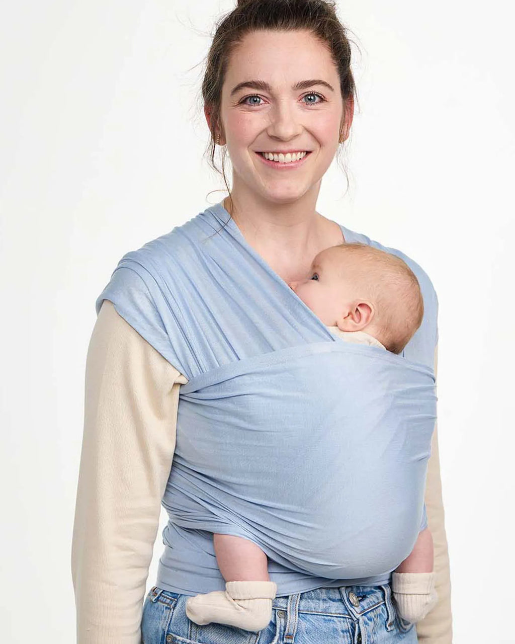 A woman wearing a light blue baby wrap with a baby inside, photographed against a clean white studio background.