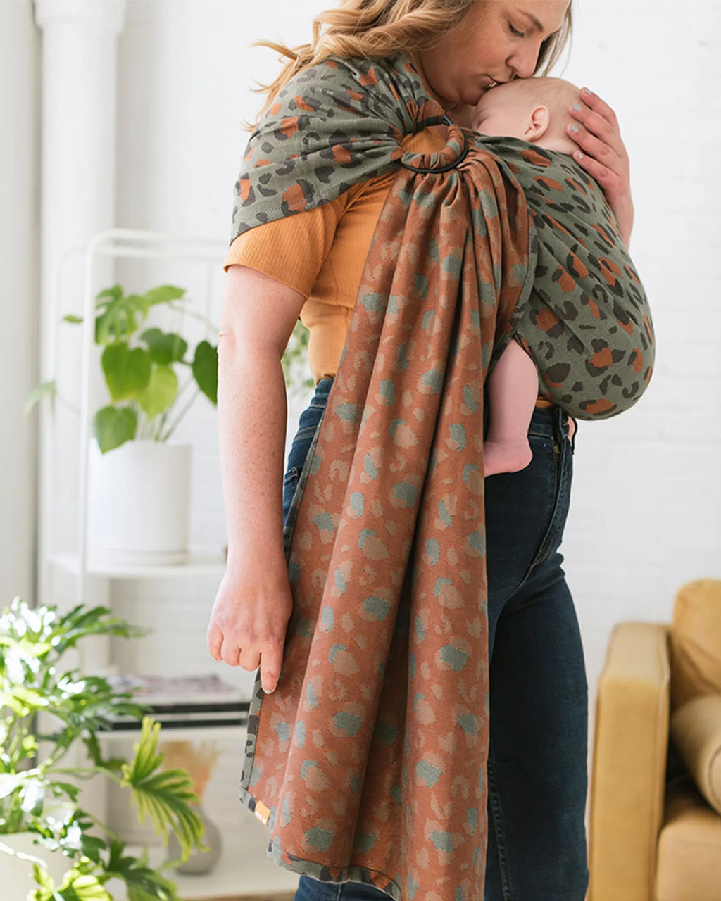 A woman wearing a baby wrap carrier with a baby, shown in a bright indoor room. The wrap features a two-tone pattern: one side is olive green with orange spots, and the other side is rust brown with light blue spots.